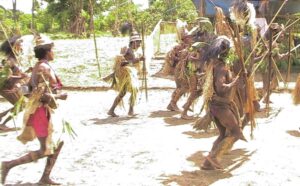 A group of national dancers from Papua, Indonesia, wear traditional tribal clothing while dancing on what appears to be sand.
