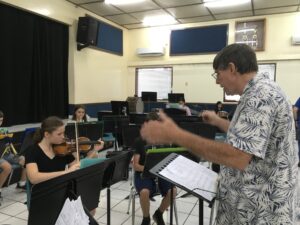 A blurry and tight shot in a school band room shows a man in a tropical floral shirt waving his arms in front of a music stand with sheet music on it - indicating he's a school band director - and various students behind music stands presumably playing instruments. One student, a woman dressed in black, is playing a violin.