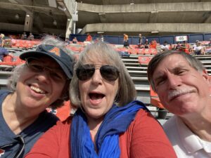 Three people face the camera in the stands of what appears to be a sports stadium. The woman on the left has short gray hair and wears a dark blue or black shirt and a blue visor with the orange letter "A"; the middle woman has shoulder-length, graying hair and wears sunglasses, a blue scarf, and a red shirt; the man on the right has short dark hair and a mustache and wears what appears to be a white polo shirt.