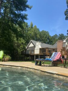 A photo taken from a backyard pool captures a light-colored house with a wooden deck surrounded by towering pine trees. Next to the pool, on the stone deck, is a lounge chair with a colorful striped towel draped over it. The sky is a cloudless, bright blue and the sun shimmers on the water of the pool.