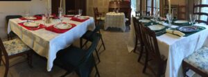 A wide-angle photo shows three tables set up for Christmas dinner in what appears to be someone's home. All are set with white tablecloths and festive dinnerware. 