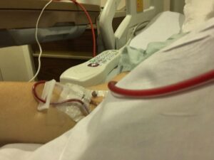 A close-up photo of a woman's arm shows blood being transfused through an IV. She's in a hospital bed, and her torso appears to be covered by a white sheet.