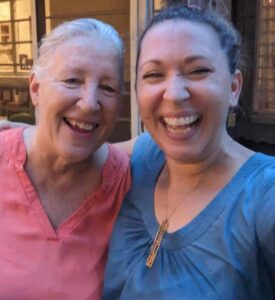 Two women smile broadly in a close-up selfie-style photo. They are sweating so it appears to be hot out. The woman on the left, who is older than the woman on the right, is wearing a pink blouse, and the woman on the right a blue blouse. 