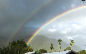 A vibrant double rainbow arcs through the sky above a tropical landscape of lush mountains and palm trees.