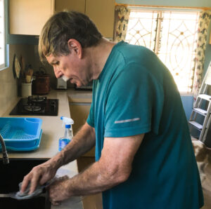 A man stands in front of a kitchen sink and appears to be scrubbing something with a rag. He's in his kitchen at home, and daylight is coming through the window in the background. He's wearing a teal-colored shirt and looking down intently.