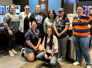 Eight 911 operators from Texas pose for a group photo in their office. Six are standing and two are kneeling in front of them. All are women. 