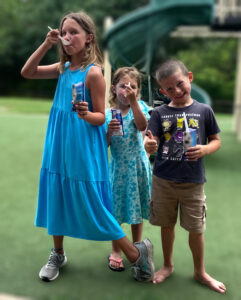 Three young children — two girls and a boy — pose for a photo while holding cups of ice cream. The two girls are wearing dresses and appear mid-bite, with their spoons in their mouths. The boy is giving a thumbs-up. They're standing on a green lawn, and a green spiral slide is behind them.