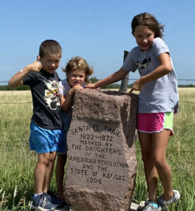 Three children stand around a stone marker that comes up the chest of the boy on the left, the chin on a girl in the middle, and just above the hips on a girl on the right. We see a couple of lines of a wire fence behind them and a background of green grass under a blue sky.