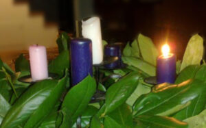 A close-up photo of an Advent wreath shows four candles sticking out from the leaves. Two candles are blue, one is white, and one is pink. One of the blue candles has been lit.