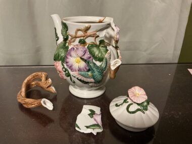 A white ceramic coffeepot accentuated with colorful flowers and leaves sits atop a wooden table in four pieces. Most of the pot is intact, but one remaining shard rests between the lid and the handle, which is shaped like a stem. The author is using the art of kintsugi to repair the broken pot, and a few cracks dusted with gold are visible.