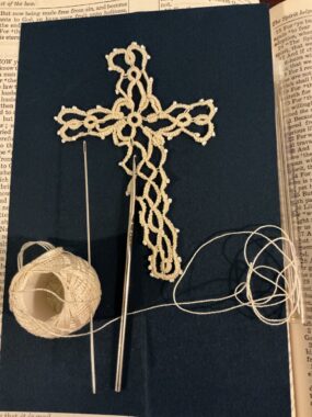 A bookmark in the shape of a cross is shown, alongside a spool of thread and needles.